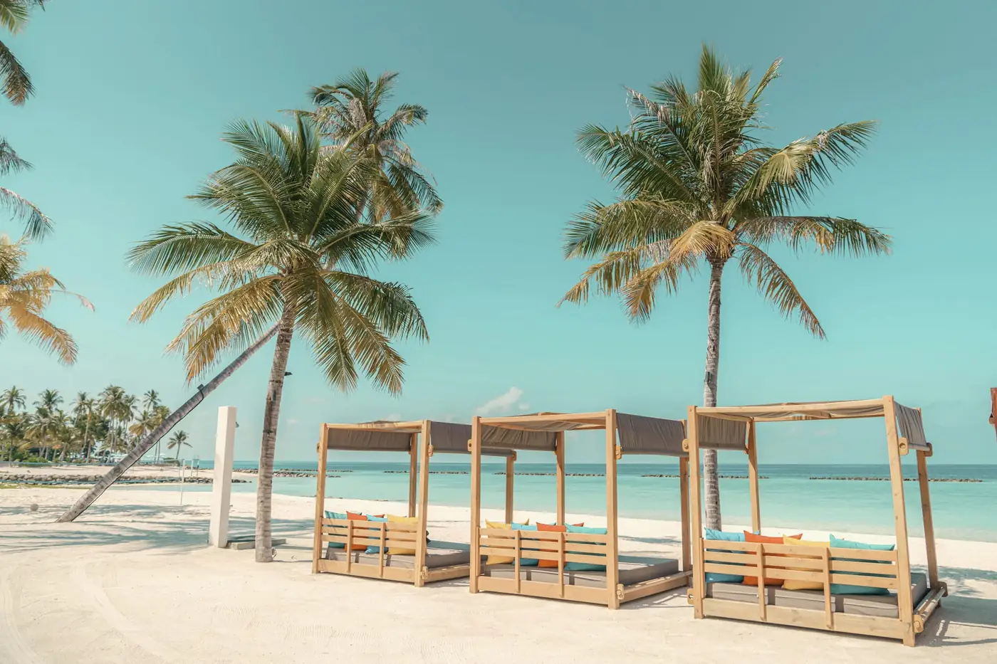Timber cabanas under two palms on a white beach beside turquoise water