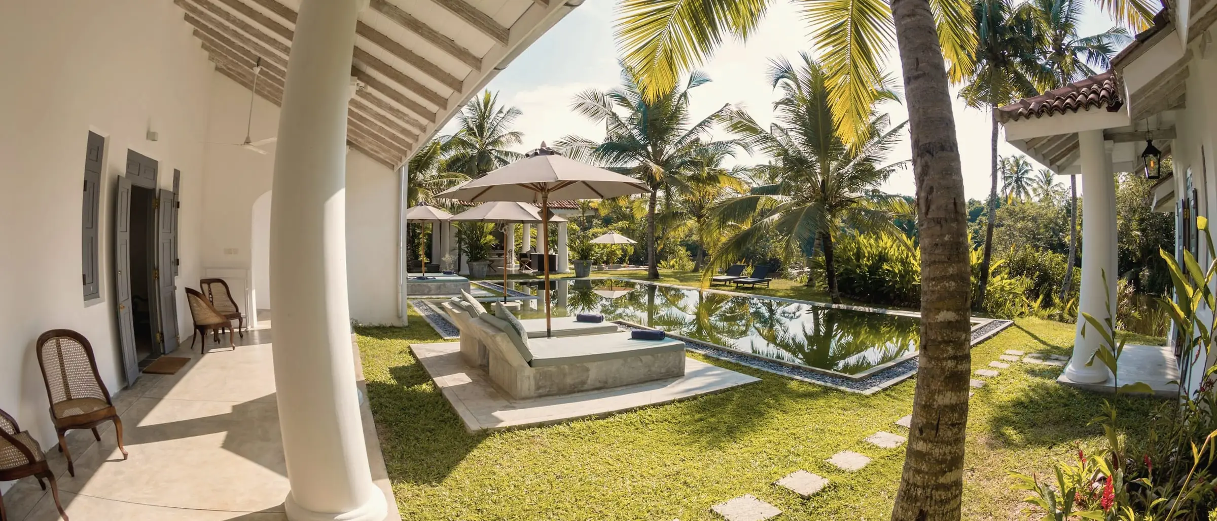 Whitewashed veranda with white columns, opening onto a lawn, palms and a long pool